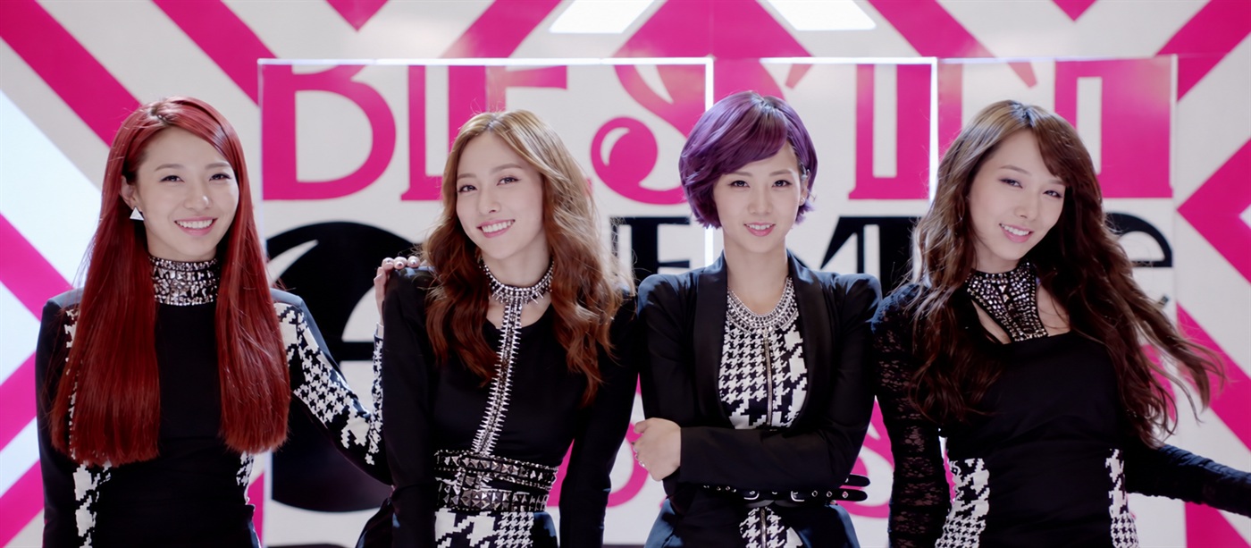The four members of BESTie in coordinated black-and-white outfits against a pink-and-white backdrop
