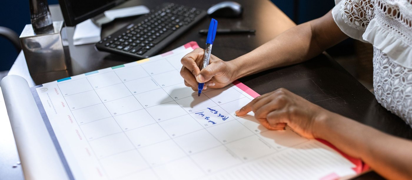 Hands writing dates into a paper monthly calendar at a desk