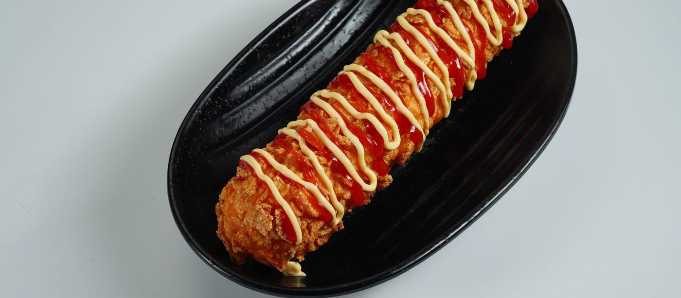 A breaded Korean corn dog zigzagged with ketchup and a pale yellow sauce on a black plate