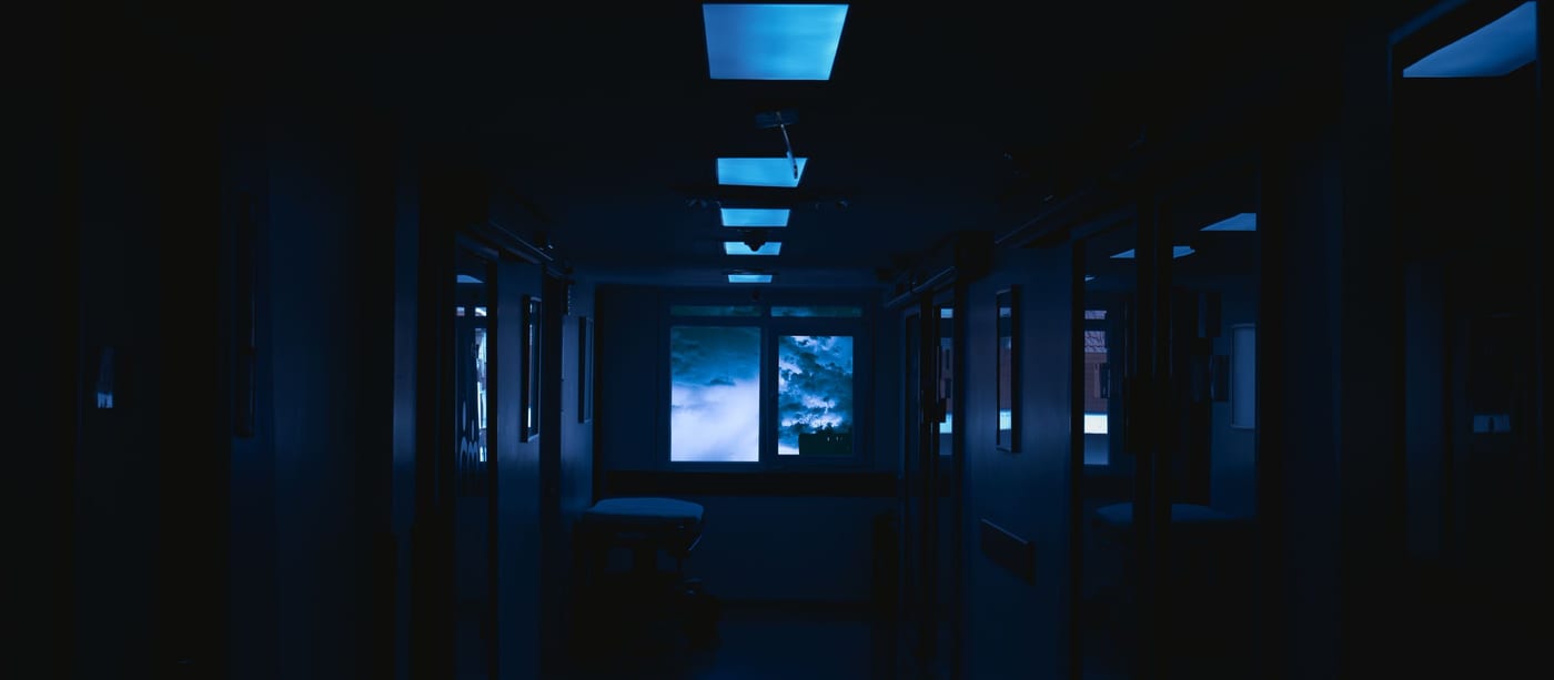 A dark hospital-like corridor lit by cold blue ceiling panels, storm clouds in the far window