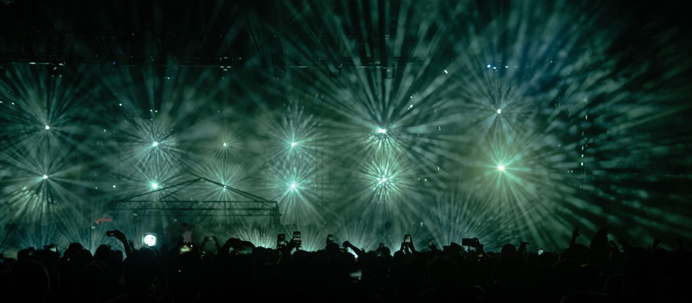 Green stage light beams bursting over a crowd of raised phones at an arena show