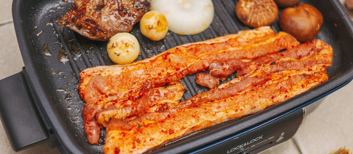 Strips of pork belly and whole garlic cloves grilling on a tabletop electric grill